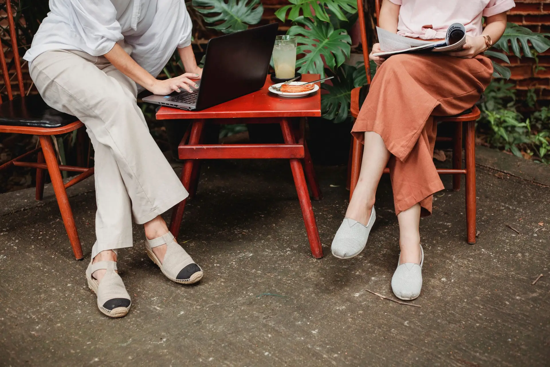 Two women at a garden table. One is typing on a laptop, the other is reading a book.